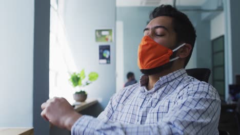 hombre de oriente medio con máscara facial usando un teléfono inteligente estornudando en su codo en una oficina moderna