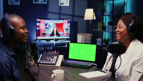 Laptop-Mit-Grünem-Bildschirm-Neben-Moderator-Und-Gast-Der-Vlogger-Show,-Die-Sich-Im-Mit-Neonlichtern-Verzierten-Wohnzimmer-Unterhalten