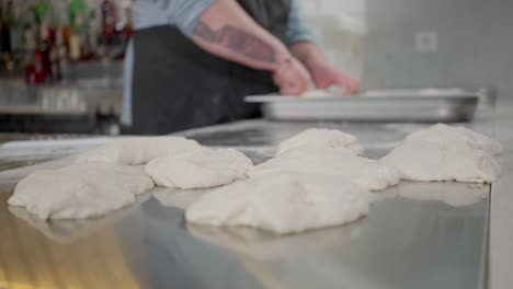 close-up of raw dough circles lying on kitchen table with blurred unrecognizable tattooed plus-size man forming pastry at background. pizza chef working in pizzeria kitchen in restaurant.