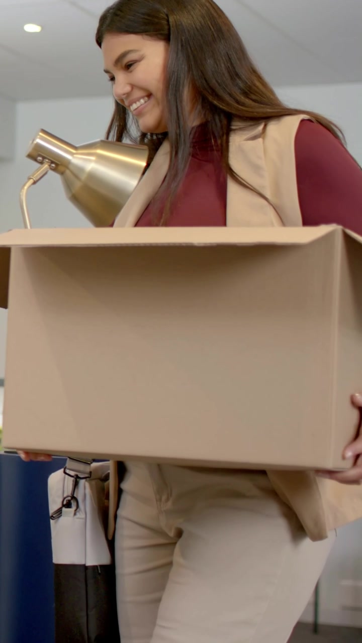 Video verticale: Donna che entra con un top color bordeaux, trasportando una scatola con una lampada dorata per allestire una scrivania in ufficio.