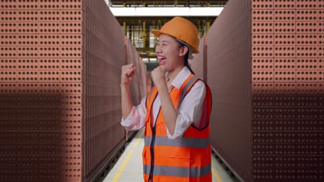 Side View Of Asian Female Engineer With Safety Helmet Screaming Goal Celebrating Working With Red Brick Packed in Stacks Are Stored