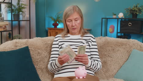 mature woman placing money into piggy bank on sofa showing savings financial responsibility at home