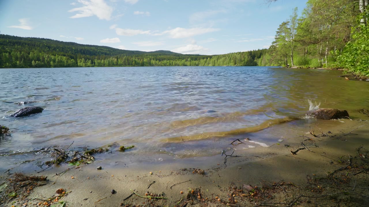 Wellen rollen sanft an einen Sandstrand, umgeben von üppigem Grün und fernen Hügeln in Norwegen. Ideal für Naturdokumentationen oder Reiseinhalte.
