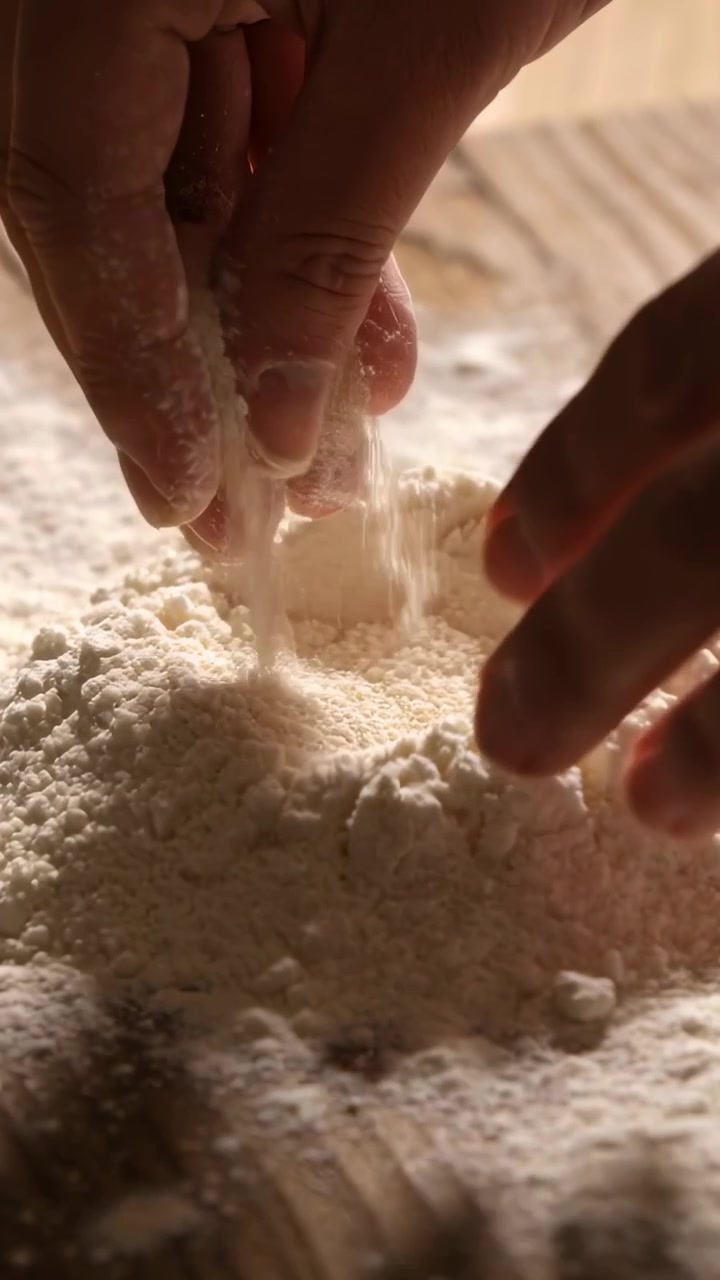Video verticale: Dita con anello che premono creando un incavo nella farina sull'asse da cucina, preparando l'impasto.