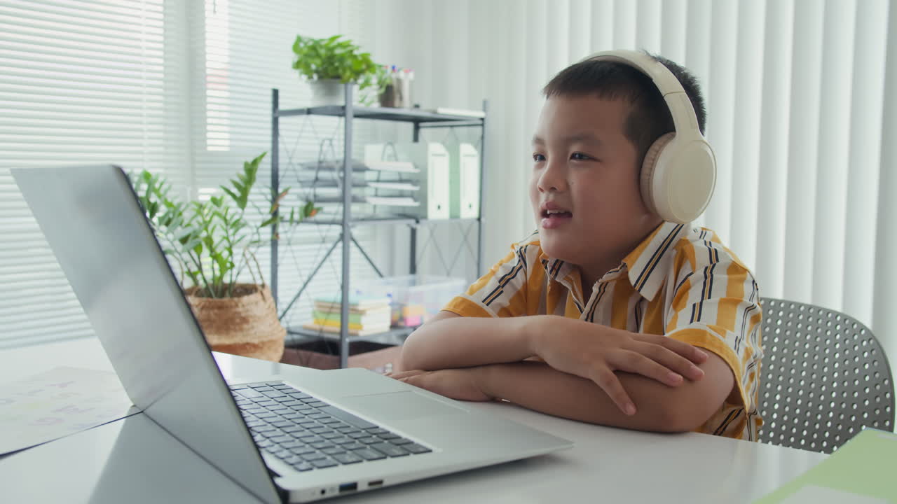 Asian Child in Headphones Talking to Teacher via Video on Laptop
