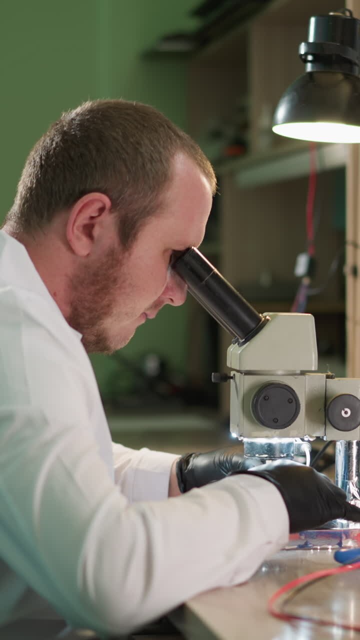 A close-up view of a technician meticulously working on an electronic component under a microscope in a laboratory, the workspace is equipped with a desk lamp and various tools