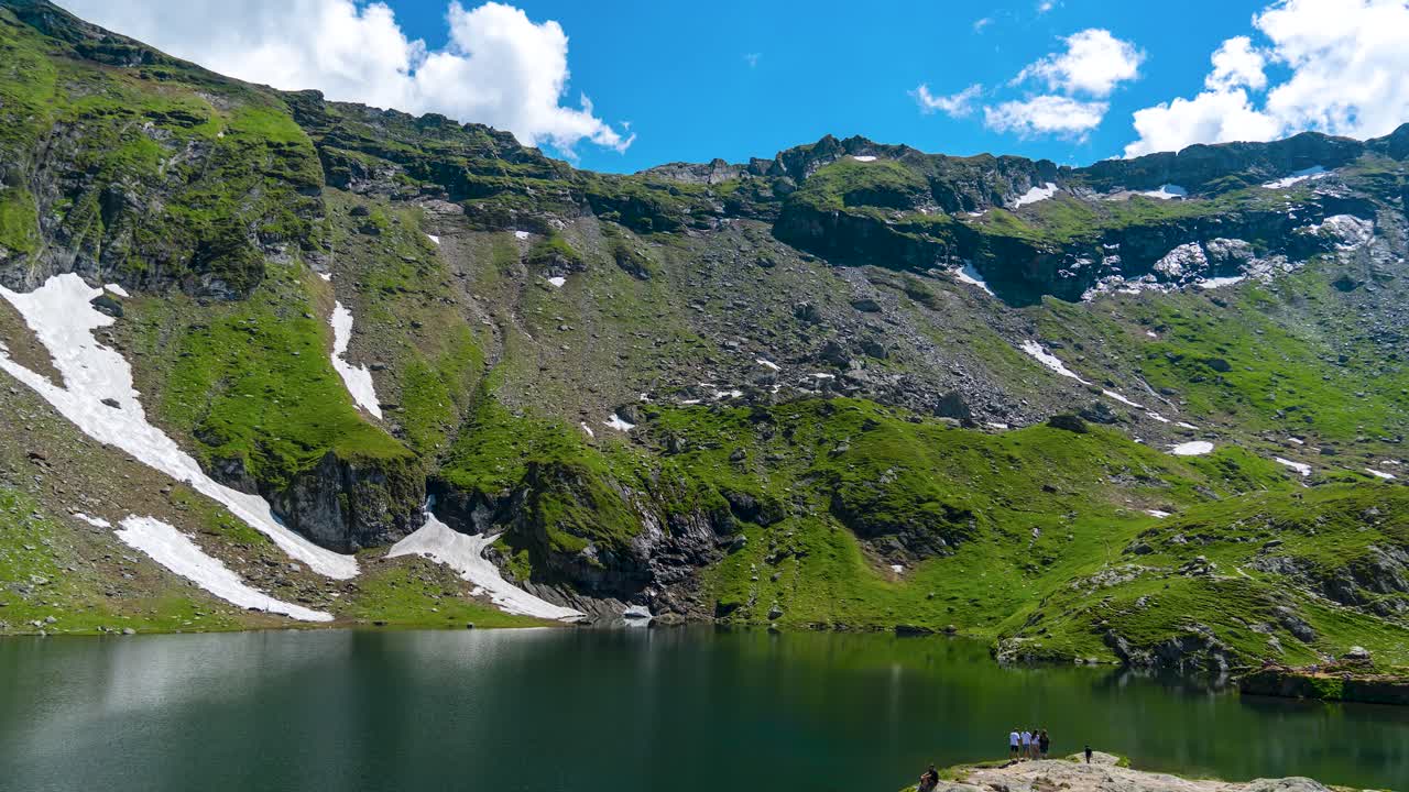 Spectacular Time-Lapse of Balea Lake A Captivating View of Snowy Mountain Peaks, Verdant Green Grass, and Clear Blue Skies with Fast-Moving White Clouds in Transylvania's Transfagarasan, Romania