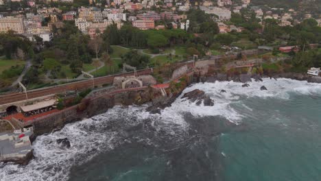 Nervi-coastline-in-genoa,-italy,-with-waves-crashing-on-rugged-rocks,-train-tracks-parallel-to-lush-gardens,-aerial-view