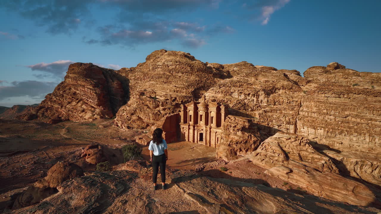 cinemagraph bucle de vídeo sin fisuras de una mujer joven en petra jordania, mirando el histórico sitio del patrimonio de la unesco ad deir monasterio tallado en piedra arenisca con el pelo en movimiento en una pintoresca tarde soleada