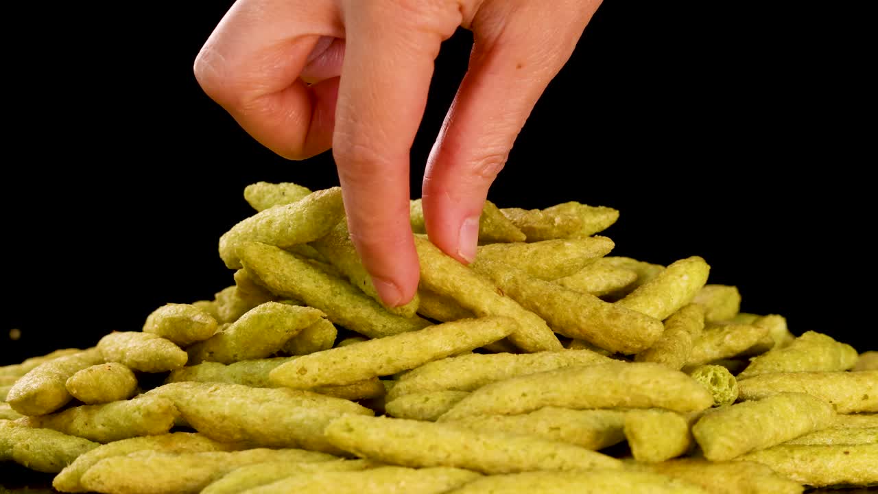 Human hand selects and lifts green pea puffed snack from pile, studio lighting, black background