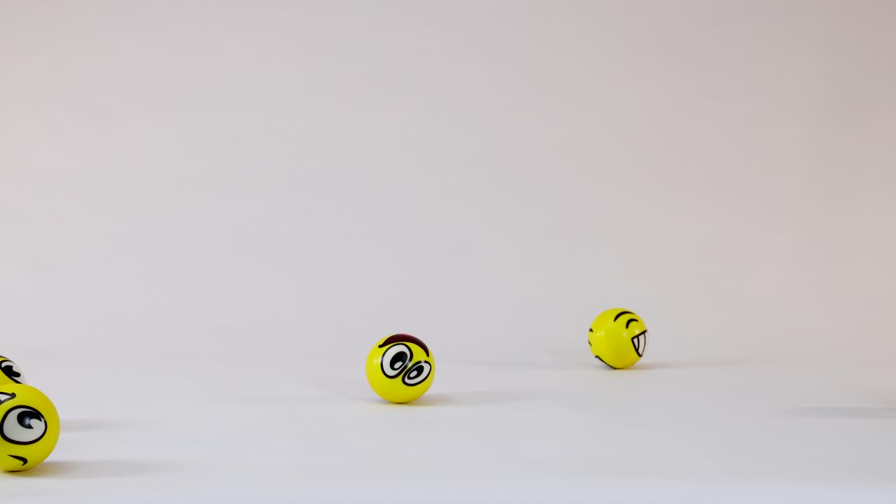 Yellow balls with various emoji faces fall, bounce, and roll across a white studio floor
