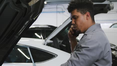 mecánico de automóviles afroamericano mirando un motor de automóvil abierto y hablando en un teléfono inteligente