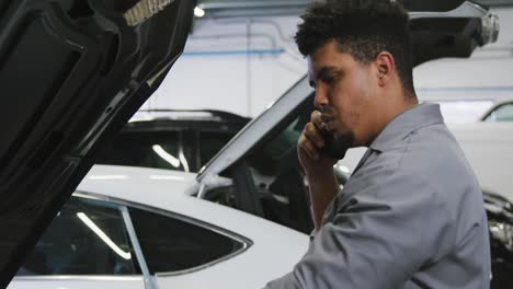 african american male car mechanic looking at an open car engine and talking on a smartphone