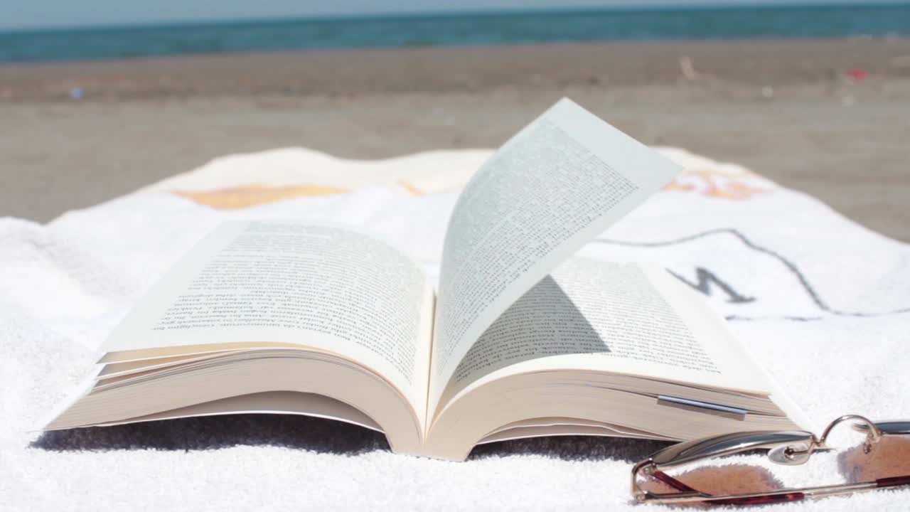 Libro aperto su un asciugamano adagiato su una spiaggia soleggiata, il vento sfoglia le pagine