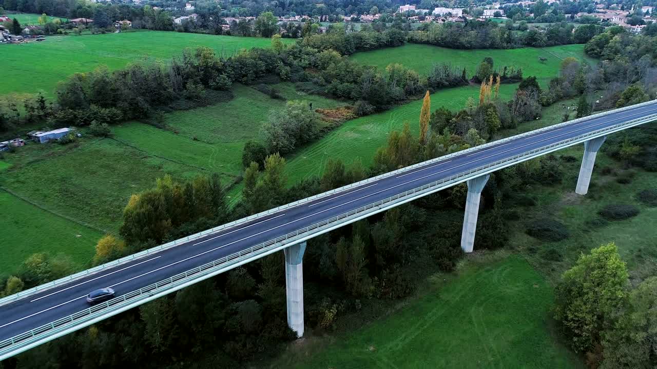 프랑스의 한 다리 위의 고속도로 위의 공중 풍경: 도로 주변의 잔디과 나무들, 빛 아래 도로를 달리는 자동차와 트럭들.