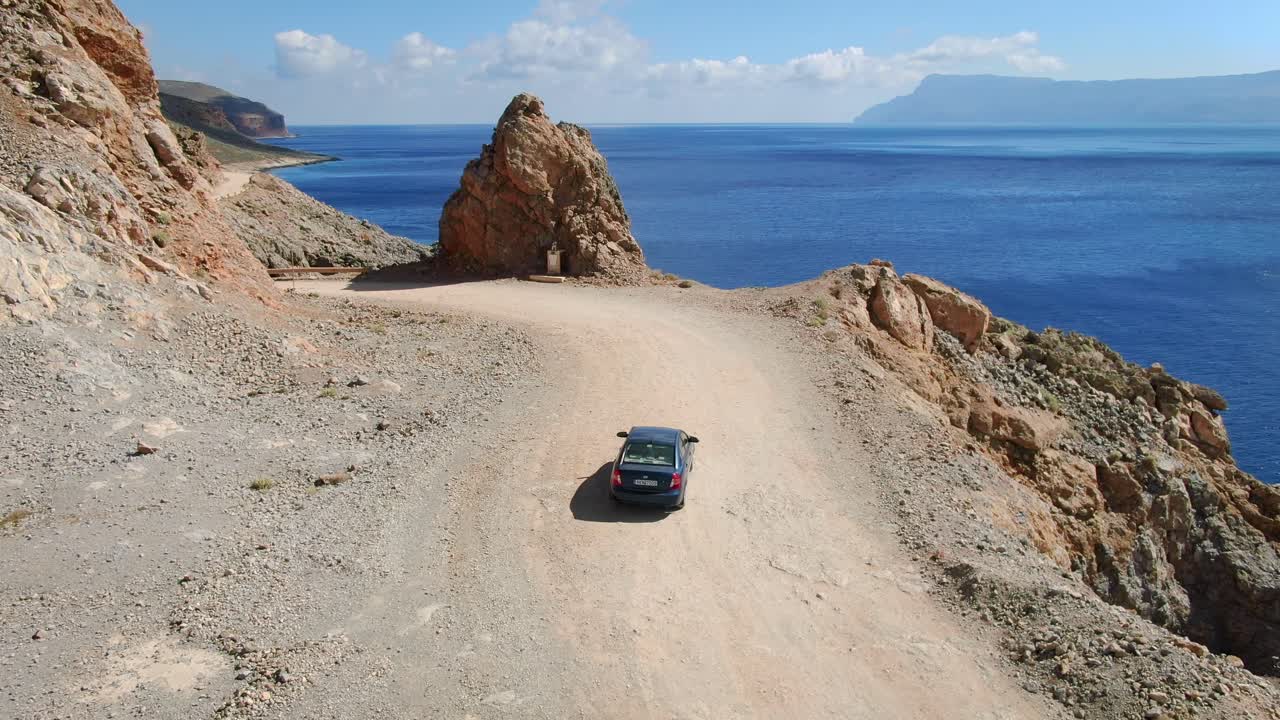A car drives slowly along a dusty road overlooking the sea on the Greek island of Crete