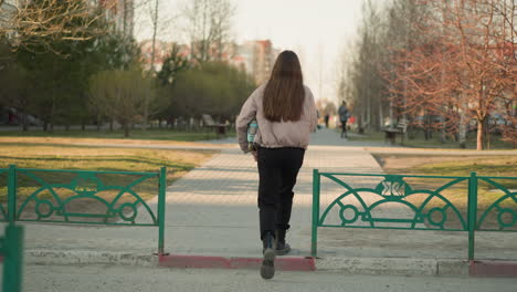 back shot of a girl in a peach jacket and black pants, holding a turquoise skateboard and walking down a park pathway in an urban park, with her long brown hair flowing, on a sunny day in early spring