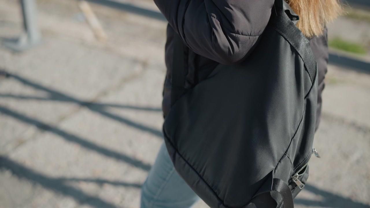 Woman carries black backpack while walking outdoors on gravel ground with metal posts in background. focus remains consistently on bag and her lower body capturing subtle movements as woman