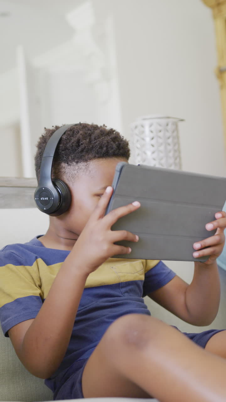 Vertical video of african american boy sitting on sofa and using tablet, in slow motion