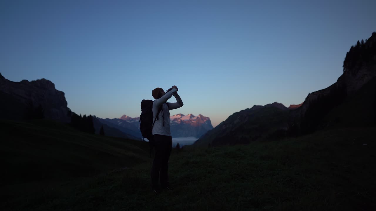 Escursionista di lungo corso che beve acqua durante un'escursione in solitaria nelle Alpi Svizzere lungo un percorso della Via Alpina. Autentico momento all'aperto che mostra resistenza, idratazione, escursionismo a lunga distanza, natura alpina, viaggio avventura.