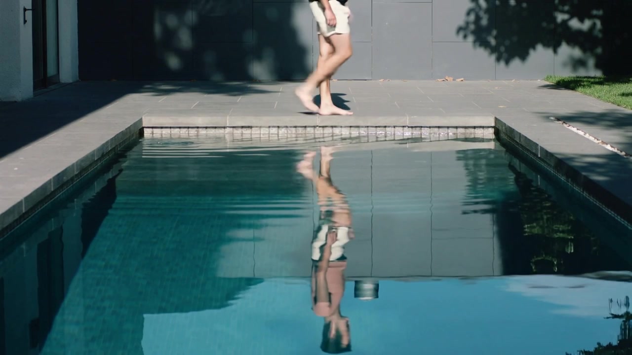 Entering frame, walking man in shorts and top crossing pool coping toward lawn, casting reflection