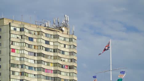 Flagge-Des-Vereinigten-Königreichs-Neben-Einem-Turmblock-Mit-Bunten-Fenstern-Und-Satelliten-Auf-Der-Spitze-Des-Gebäudes-Sonniger-Tag-Zeitlupe
