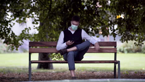 An-elegant-young-man-in-a-park-wearing-a-black-waistcoat-and-white-shirt-and-a-green-protective-Covid-19-mask,-sitting-on-a-park-bench-beside-a-fountain,-picking-up-a-business-call,-static-4k