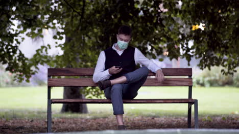 Un-Joven-Elegante-En-Un-Parque-Con-Un-Chaleco-Negro-Y-Una-Camisa-Blanca-Y-Una-Máscara-Protectora-Verde-Covid-19,-Sentado-En-Un-Banco-Del-Parque-Junto-A-Una-Fuente,-Atendiendo-Una-Llamada-De-Negocios,-Estático-4k