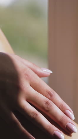 cu shot, slow motion: young lady strokes long bare leg by manicured hand sitting near window at home in early sunny morning extreme close view