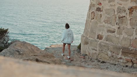 Reveal-camera-shot-of-young-woman-walking-by-the-old-fort-near-the-sea-ocean-water