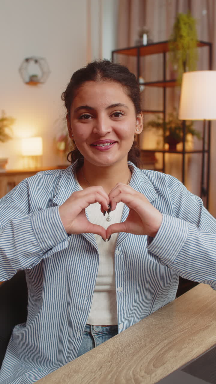 Happy indian young woman makes symbol of love showing heart sign to camera at home office desk