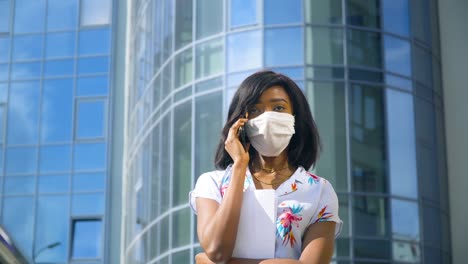 african american young business woman in protective mask is talking on phone outside near modern office building. end of quarantine. stop virus