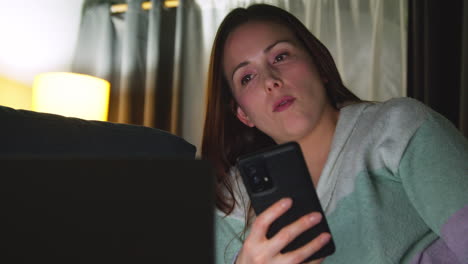 Close-Up-Of-Woman-Sitting-On-Sofa-At-Home-At-Night-Streaming-Or-Watching-Movie-Or-Show-On-Laptop-And-Scrolling-Internet-On-Phone