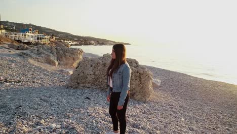 caucasian woman standing on rocky beach by ocean and bright white sun on horizon, kafarabida, lebanon, spinning aerial