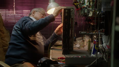 Elderly-caucasian-man-fixes-radio-equipment-in-dark-room,-side-view