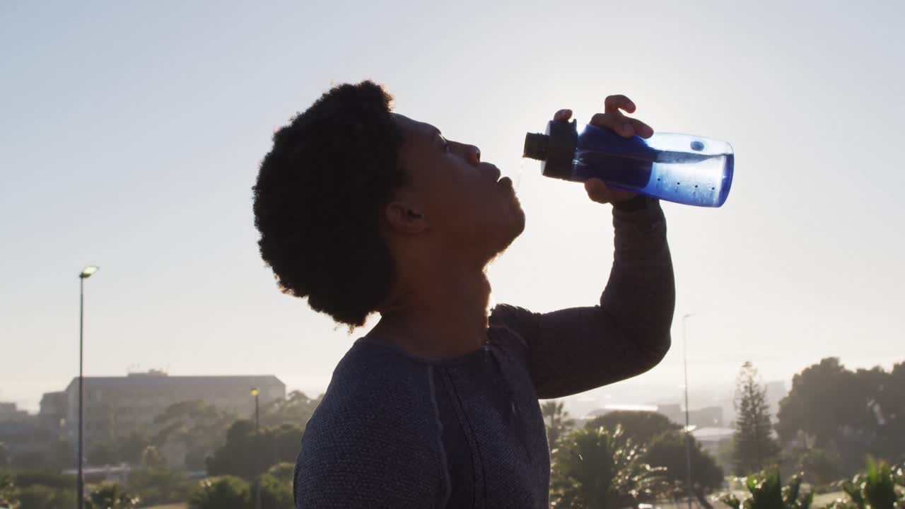 Homme afro-américain sportif s'entraînant en plein air en ville, se reposant et buvant à sa bouteille d'eau.