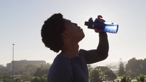fit african american man exercising outdoors in city, resting and drinking from water bottle