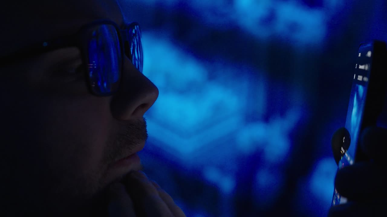 Primer plano cinemático de un hombre con gafas viendo contenido en streaming en un smartphone por la noche bajo luz azul, representando el uso nocturno de pantallas y el estilo de vida digital.