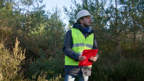 male engineer looking around the trees at the forest while holding on to his clipboard, handheld