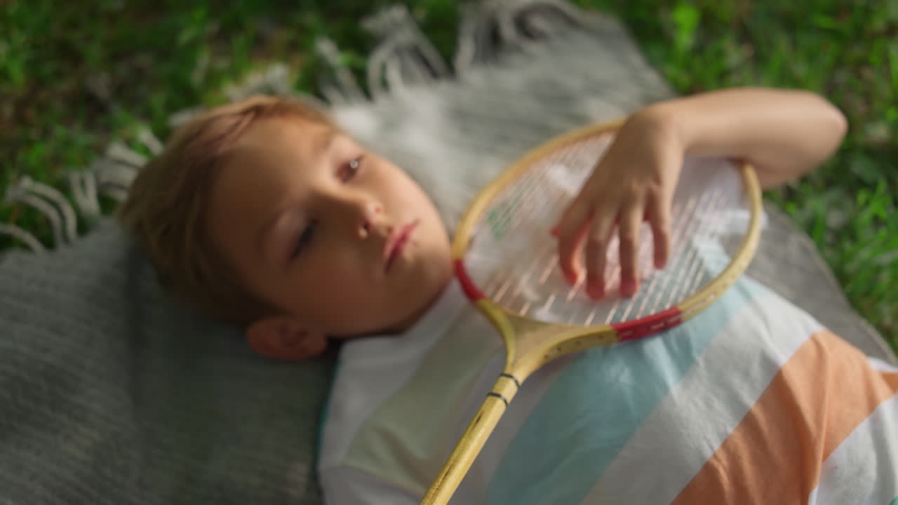 Closeup tired boy lying blanket in summer park examining badminton racket net.