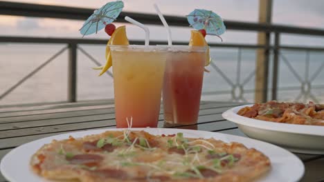 pizza, pasta and cocktail drinks on a table along the beach during a peaceful sunset