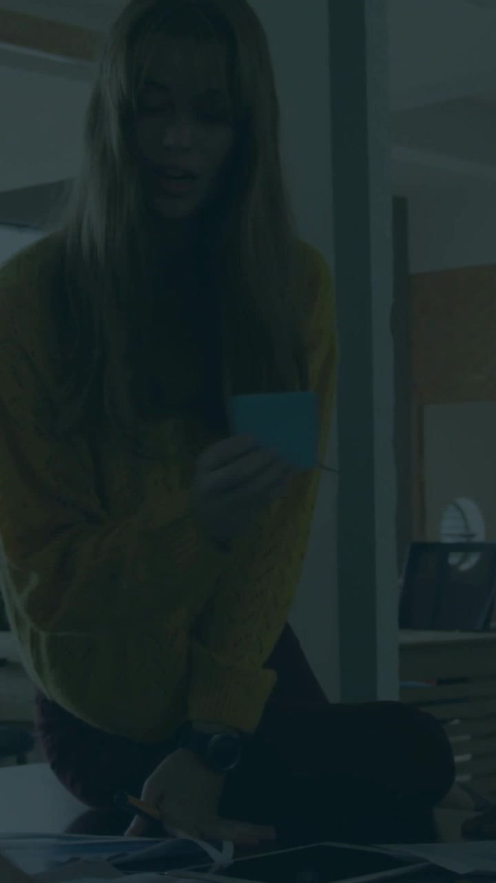 Donna in maglione giallo che si protende e consegna una carta azzurra a una scrivania in ufficio, con sovrapposizione verticale.