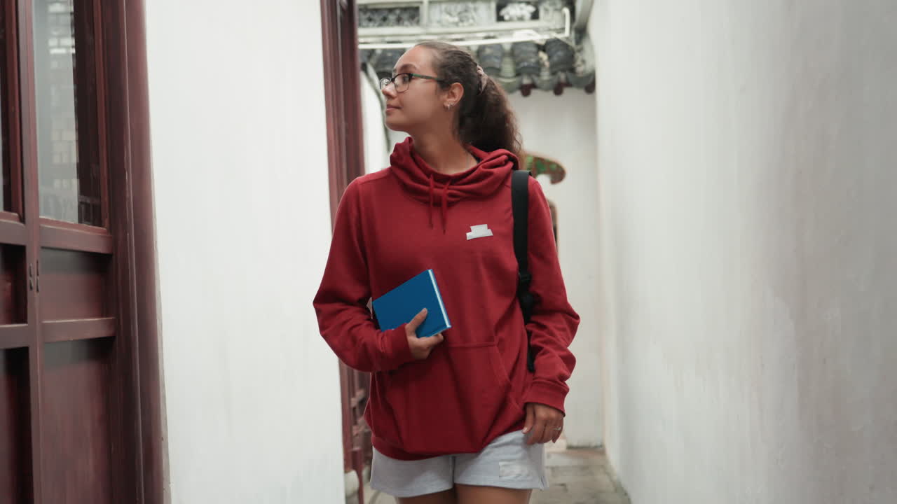Jovem estudante latina com caderno caminhando pela entrada de um pátio histórico, vestindo moletom vermelho e segurando um diário azul enquanto observa detalhes arquitetónicos e lanternas penduradas, estudo espontâneo