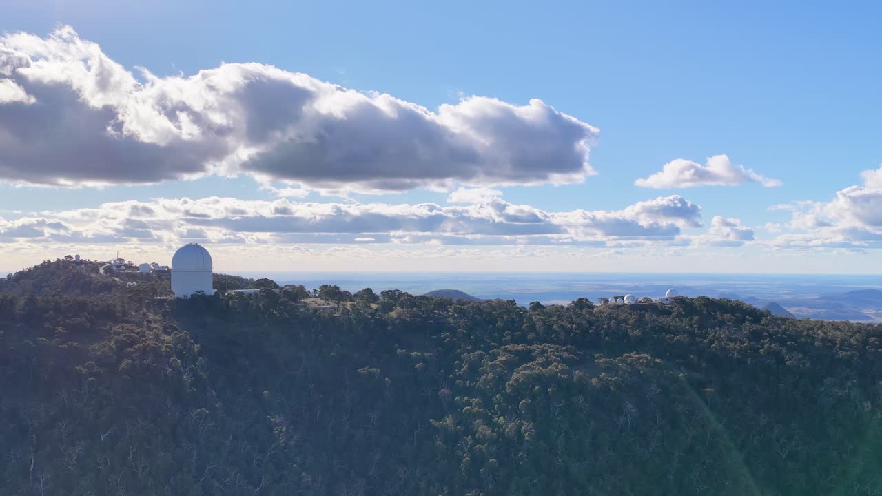 Drone footage smoothly pans across a large white observatory dome atop a forested mountain ridge under bright daylight, with scattered clouds and expansive sky