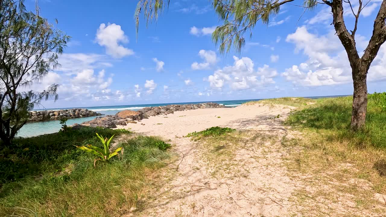 Camera moves along sandy path through greenery toward blue ocean under bright daylight sky