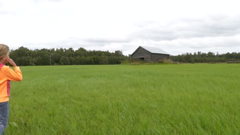 freedom of summer break, alone blond young girl walking on grass field, approaching old barn, slow motion