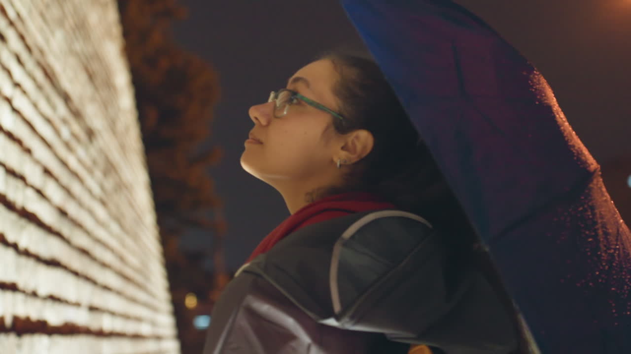 Mulher asiática com guarda-chuva azul olhando para a parede, mochila visível, luzes da cidade e bokeh ao fundo, pingos de chuva suaves na cobertura, humor reflexivo e esperançoso, viajante pausando para absorver a paisagem noturna urbana.