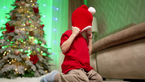 toddler kneeling near the sofa holding a santa cap on his face. child stands up and runs away. low angle view. christmas tree at backdrop.