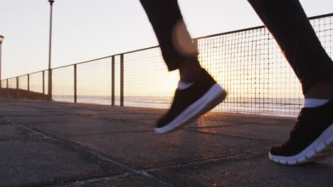 Sección-Baja-De-Un-Hombre-Afroamericano-Haciendo-Ejercicio-Al-Aire-Libre,-Corriendo-Junto-Al-Mar-Al-Atardecer