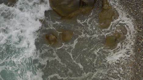 Ocean-Waves-Crash-Over-Rocky-Boulders-Along-a-Coastal-Pebbled-Beach-with-a-Top-View-in-Looc-Bay,-Philippines